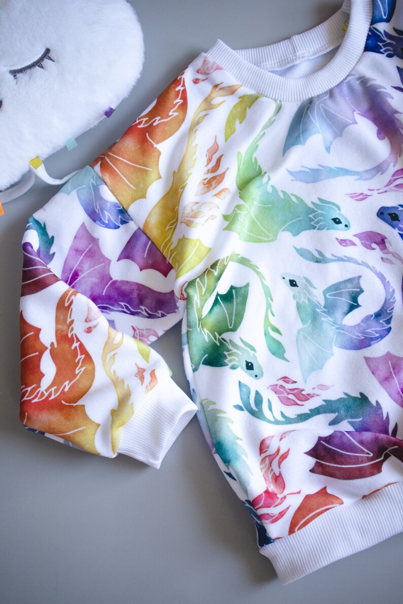 Puede incluir: Sudadera blanca con estampado de acuarela de dragones coloridos. Los dragones son en tonos de naranja, amarillo, verde, azul y morado.