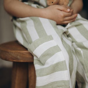 May include: A child wearing a green and white striped jumpsuit sits on a wooden stool, holding a brown stuffed bunny.