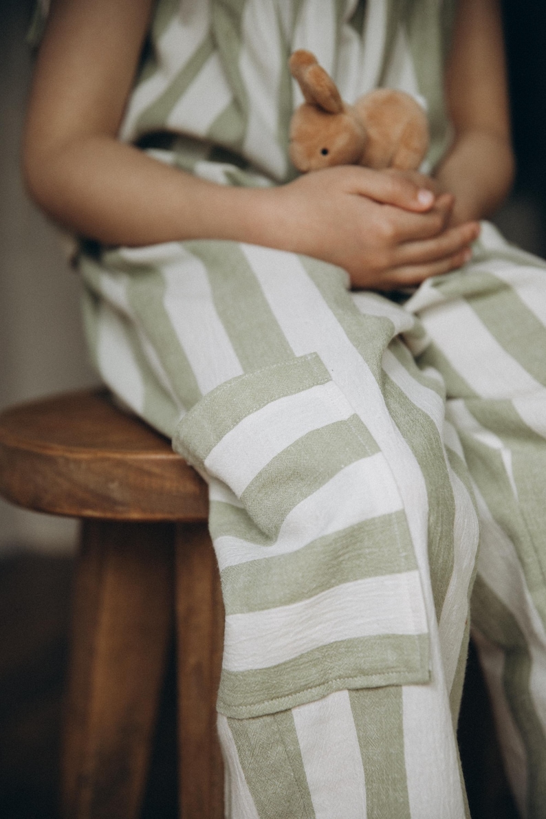 May include: A child wearing a green and white striped jumpsuit sits on a wooden stool, holding a brown stuffed bunny.
