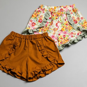 Puede incluir: Dos pares de pantalones cortos para niñas. Un par es marrón con volantes y el otro par es blanco con estampado floral y volantes verdes.