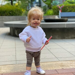 Puede incluir: Un ni&ntilde;o peque&ntilde;o con cabello rubio que lleva un su&eacute;ter blanco, pantalones marrones y zapatillas blancas. El ni&ntilde;o sostiene una pajita roja y sonr&iacute;e a la c&aacute;mara.