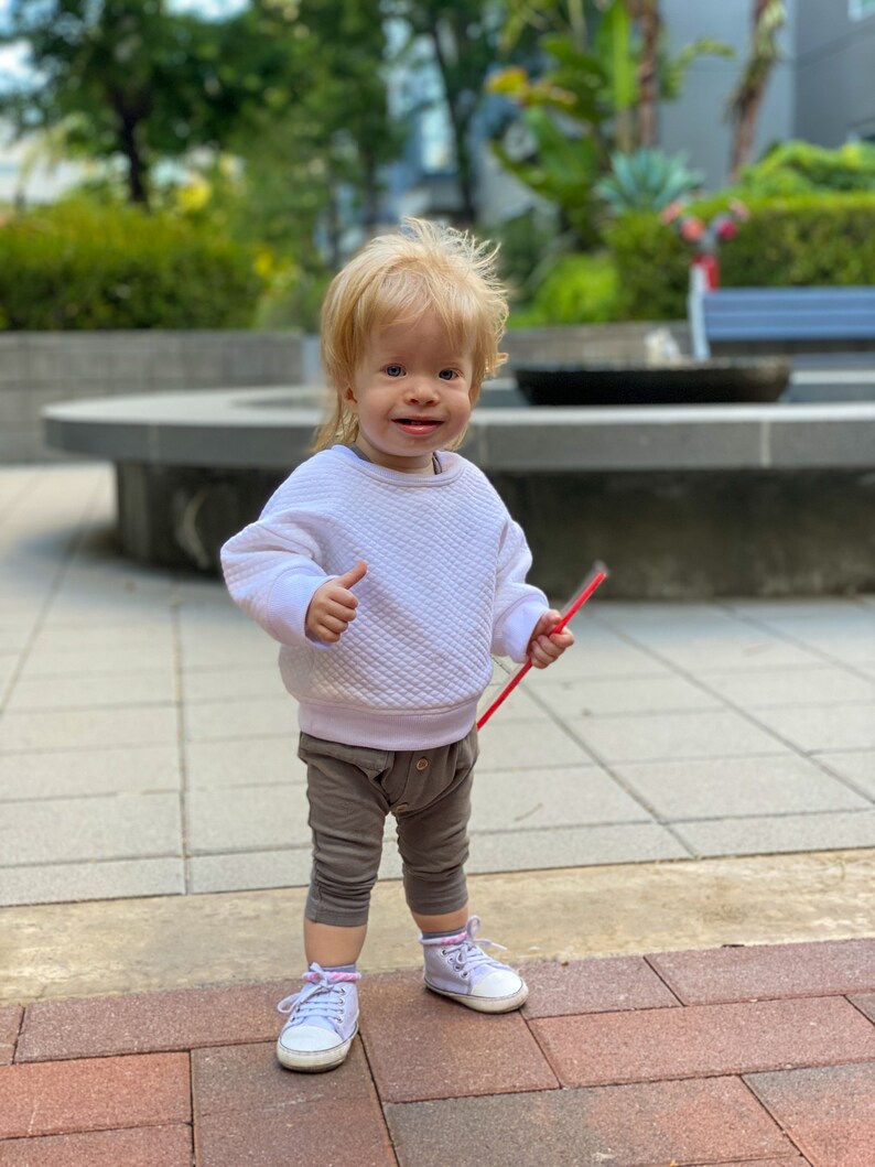 Puede incluir: Un ni&ntilde;o peque&ntilde;o con cabello rubio que lleva un su&eacute;ter blanco, pantalones marrones y zapatillas blancas. El ni&ntilde;o sostiene una pajita roja y sonr&iacute;e a la c&aacute;mara.