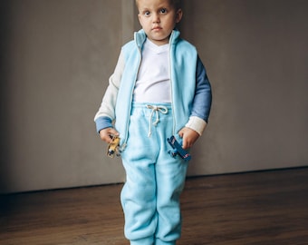 May include: A young child wearing a light blue tracksuit with a white t-shirt underneath. The tracksuit has a white and blue color block design on the sleeves. The child is holding a toy airplane in one hand.