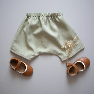 May include: A pair of light green linen shorts with a gathered waistband. The shorts are laid flat on a light gray surface with a sprig of dried flowers and two pairs of brown leather shoes with white trim.