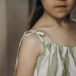 May include: A young girl wearing a green and white striped sleeveless dress with thin straps that tie at the shoulders.