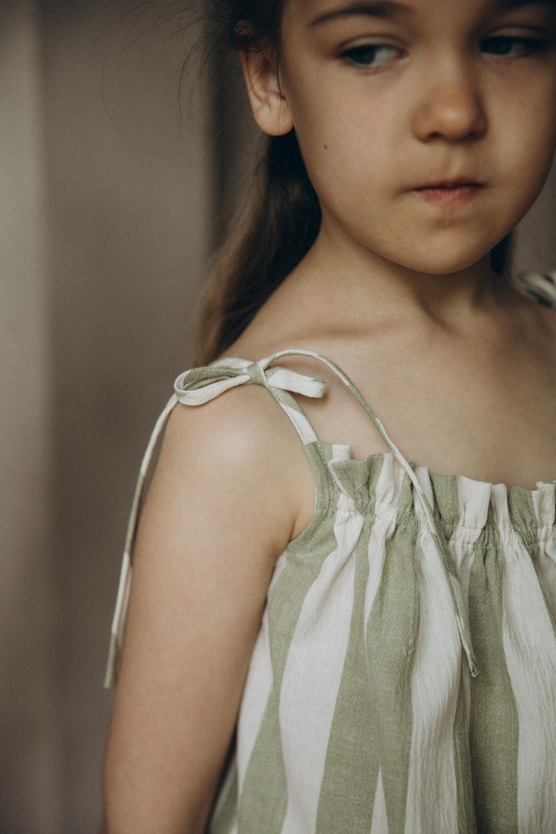 May include: A young girl wearing a green and white striped sleeveless dress with thin straps that tie at the shoulders.