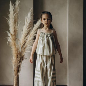 May include: A young person wearing a green and white striped top and matching pants. The outfit has a relaxed, casual style. The person is standing in front of a wall with a vase of dried pampas grass.