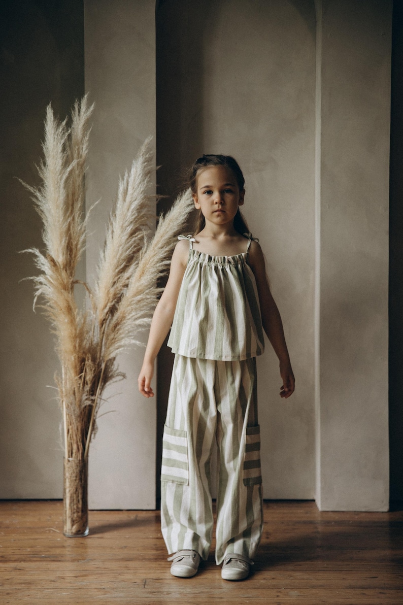 May include: A young person wearing a green and white striped top and matching pants. The outfit has a relaxed, casual style. The person is standing in front of a wall with a vase of dried pampas grass.