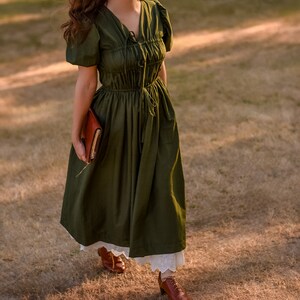 May include: A woman wearing a dark green, long-sleeved dress with a gathered bodice and a white lace trim. She is holding a brown leather journal in her left hand and is walking on a grassy field.
