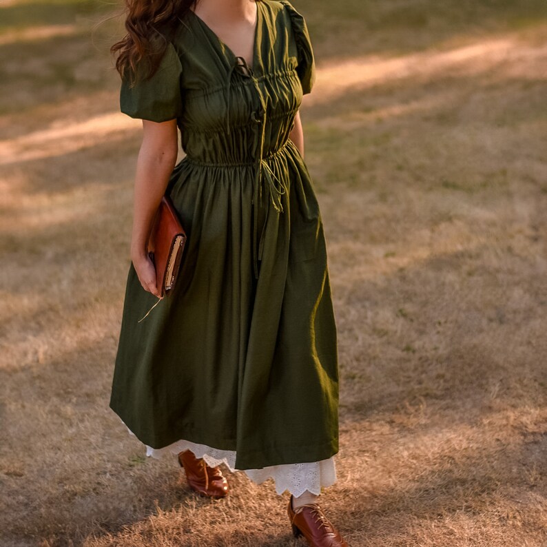 May include: A woman wearing a dark green, long-sleeved dress with a gathered bodice and a white lace trim. She is holding a brown leather journal in her left hand and is walking on a grassy field.