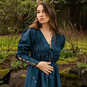 May include: A woman wearing a dark blue dress with a gathered bodice and long sleeves. The dress has a V-neckline and a tie at the waist. The sleeves have a lace-up detail at the cuffs.