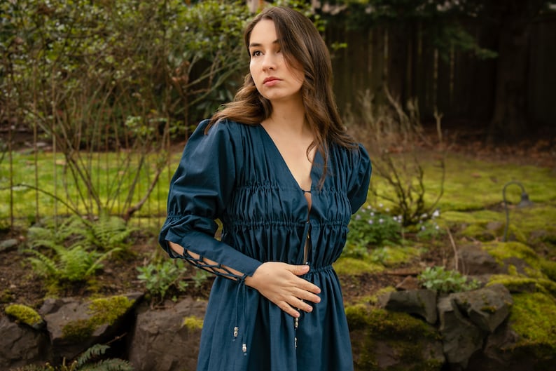 May include: A woman wearing a dark blue dress with a gathered bodice and long sleeves. The dress has a V-neckline and a tie at the waist. The sleeves have a lace-up detail at the cuffs.