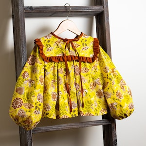 May include: A yellow blouse with a brown ruffled collar and long sleeves. The blouse has a floral pattern with brown and green accents.