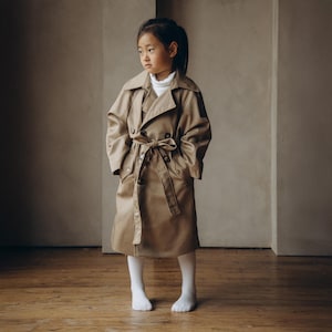 Puede incluir: Una persona joven lleva un abrigo beige de gabardina con cierre de doble botonadura y cinturón. El abrigo tiene bolsillos en la parte delantera y se lleva sobre un jersey de cuello alto blanco.