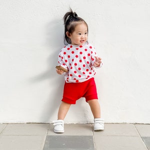Puede incluir: Un ni&ntilde;o peque&ntilde;o con una camisa blanca con lunares rojos y pantalones cortos rojos se para frente a una pared blanca. El ni&ntilde;o sostiene un peque&ntilde;o objeto marr&oacute;n en su mano.