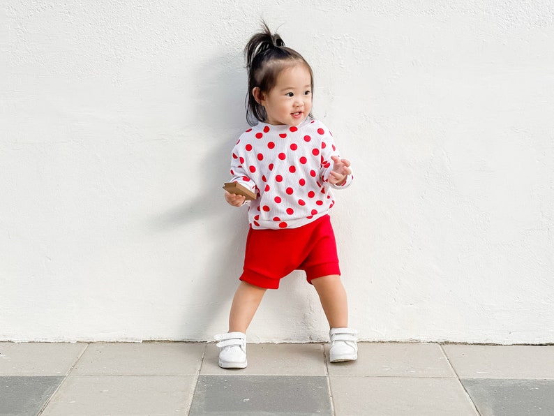 Puede incluir: Un ni&ntilde;o peque&ntilde;o con una camisa blanca con lunares rojos y pantalones cortos rojos se para frente a una pared blanca. El ni&ntilde;o sostiene un peque&ntilde;o objeto marr&oacute;n en su mano.