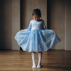 May include: A young girl wearing a light blue dress with a lace skirt and long sleeves. She is standing on a wooden floor and is looking down at the dress.