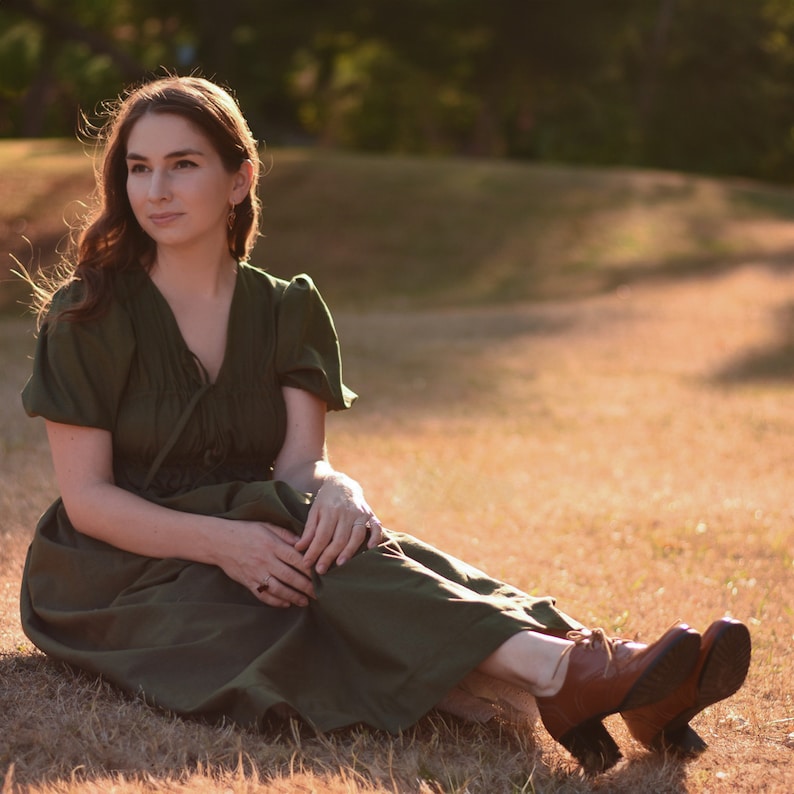 May include: A woman wearing a green dress sits in a field of tall grass. She is wearing brown shoes and has her hair pulled back. The sun is shining brightly and the grass is a golden color.