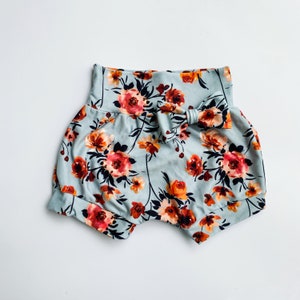 May include: Baby bloomers with a floral pattern. The shorts are light blue with a repeating pattern of orange and red flowers and dark green leaves. A matching bow is on the front of the shorts.