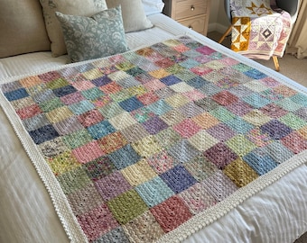 May include: A handmade crochet blanket featuring a grid of multicolored square patches in various shades of pink, blue, green, yellow, and grey. The blanket is finished with a simple white crocheted border around the entire perimeter.