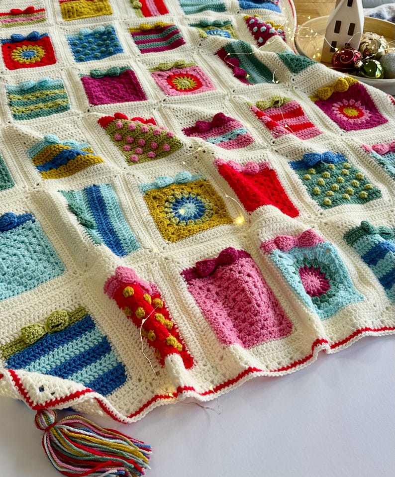 May include: A white crocheted blanket with a red border and a colorful tassel. The blanket is made up of squares with different patterns, including stripes, polka dots, and flowers. The blanket is perfect for a cozy winter night.