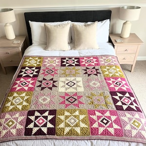 Op de afbeelding: Een handgemaakte gehaakte deken met een sterrenpatroon in roze, bordeaux, olijfgroen en crèmetinten. De deken ligt op een bed met wit beddengoed en twee beige kussens. De deken is ongeveer 200 cm x 200 cm groot.