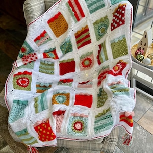 May include: A white crochet blanket with a red border and a variety of colorful squares. The squares feature different patterns, including stripes, polka dots, and flowers. The blanket is draped over a chair.