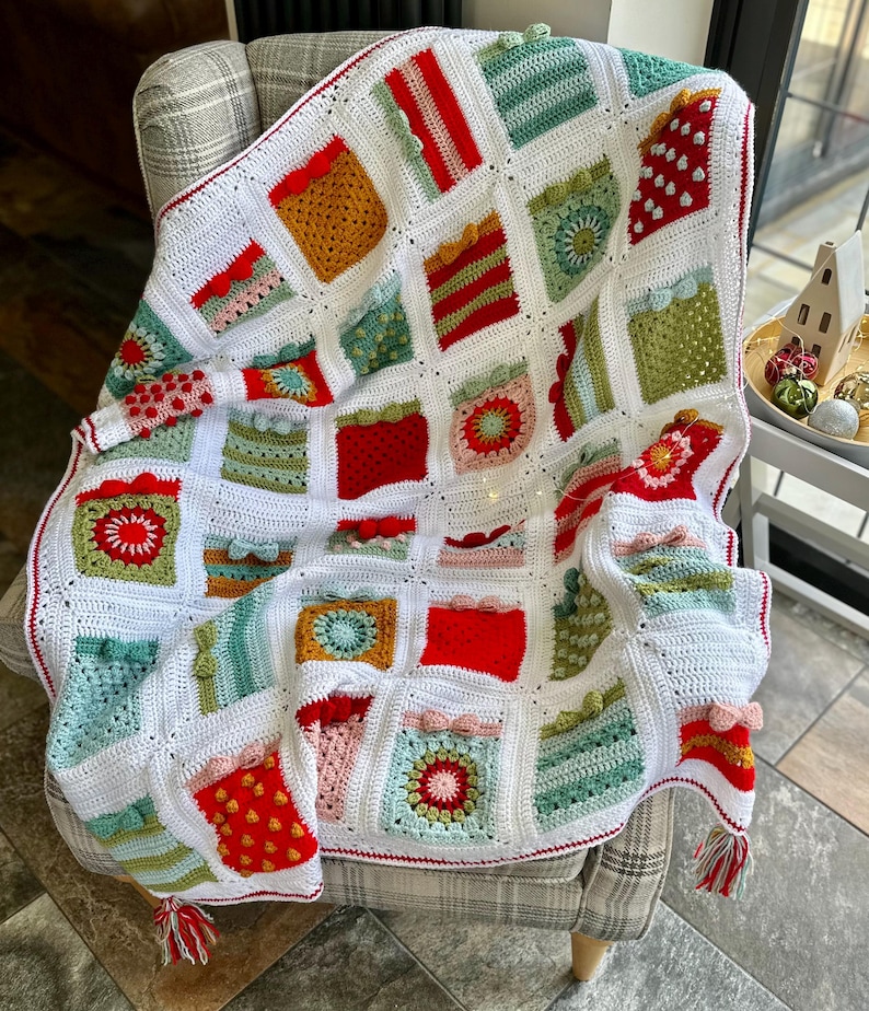 May include: A white crochet blanket with a red border and a variety of colorful squares. The squares feature different patterns, including stripes, polka dots, and flowers. The blanket is draped over a chair.