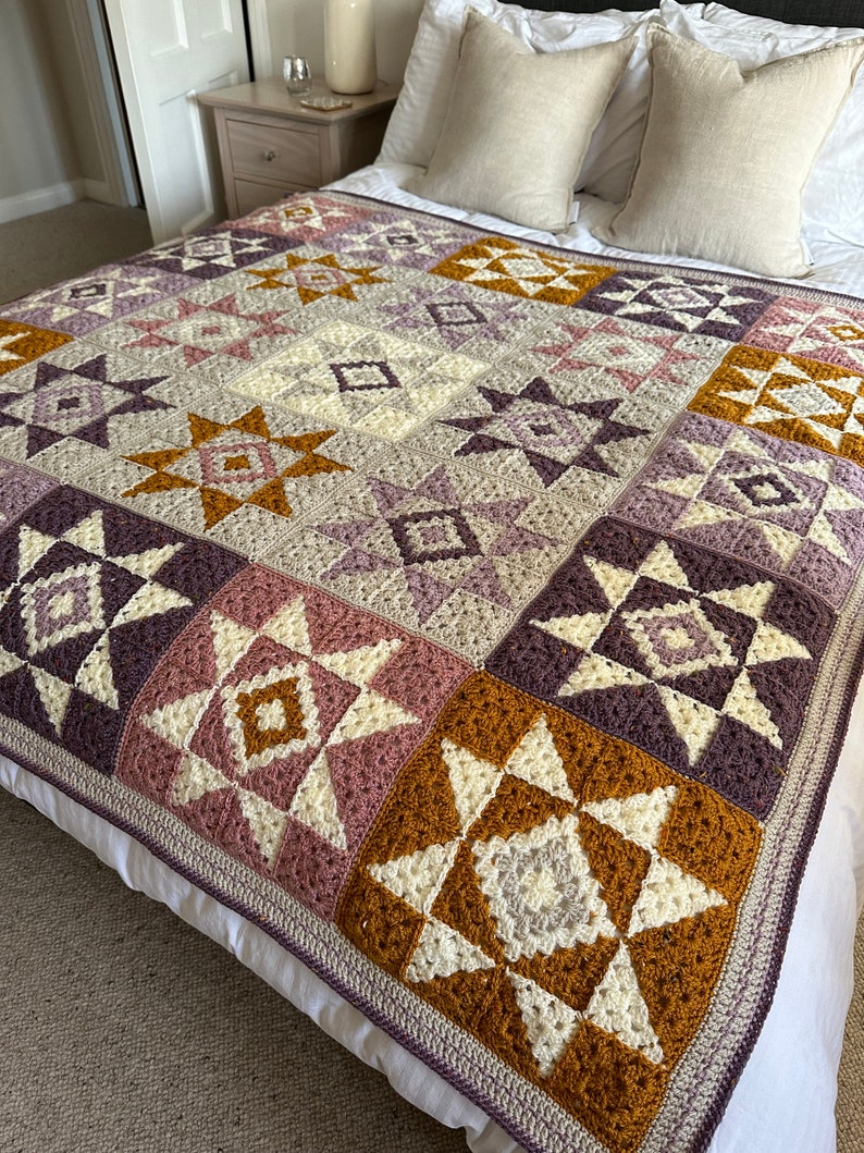Peut inclure: Une couverture en crochet avec un motif g&eacute;om&eacute;trique dans les tons de violet, rose, blanc et marron. La couverture est compos&eacute;e de carr&eacute;s avec diff&eacute;rents motifs d'&eacute;toiles et de diamants.