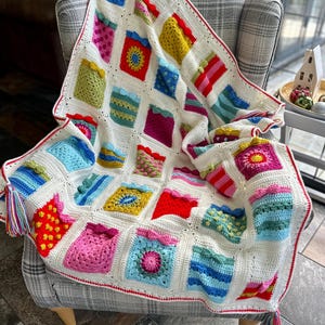 May include: A colorful crocheted blanket with a white background and a red border. The blanket is made up of squares with different patterns and colors, including pink, blue, green, yellow, and red. The blanket is folded and draped over a chair.