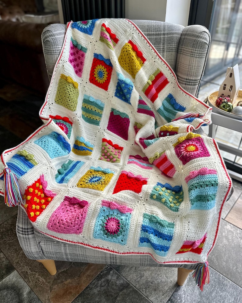May include: A colorful crocheted blanket with a white background and a red border. The blanket is made up of squares with different patterns and colors, including pink, blue, green, yellow, and red. The blanket is folded and draped over a chair.