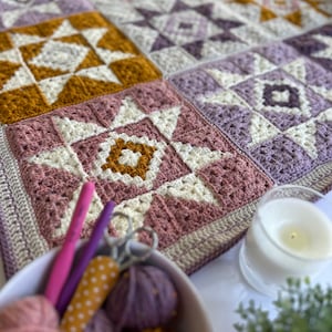 Peut inclure: Une couverture au crochet avec un motif patchwork pr&eacute;sentant diff&eacute;rentes nuances de violet, rose, blanc et jaune. La couverture est compos&eacute;e de carr&eacute;s avec diff&eacute;rents motifs d'&eacute;toiles. La couverture est pos&eacute;e sur une table avec de la laine, des aiguilles &agrave; tricoter et des ciseaux.