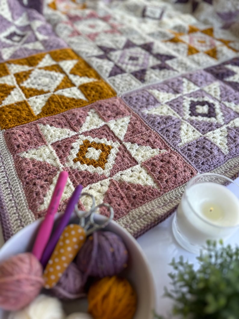 Peut inclure: Une couverture au crochet avec un motif patchwork pr&eacute;sentant diff&eacute;rentes nuances de violet, rose, blanc et jaune. La couverture est compos&eacute;e de carr&eacute;s avec diff&eacute;rents motifs d'&eacute;toiles. La couverture est pos&eacute;e sur une table avec de la laine, des aiguilles &agrave; tricoter et des ciseaux.