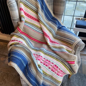 Può includere: Una coperta all'uncinetto con strisce blu, verdi, rosa e bianche. La coperta è drappeggiata su una sedia con motivo a quadri.