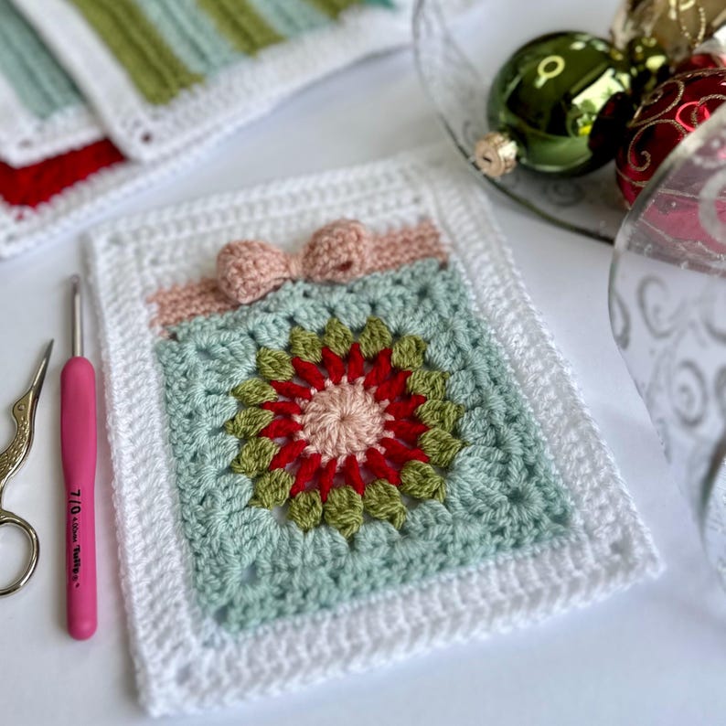 May include: A white crocheted square with a green, red, and pink flower design in the center. A pink bow is attached to the top of the square.