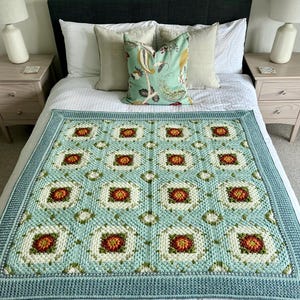 Op de afbeelding: Een handgemaakte, vierkante gehaakte deken in lichtblauw, wit en rood-oranje. De deken heeft een herhalend bloemenpatroon in vierkante blokken, omzoomd door een effen lichtblauwe rand. De deken wordt gepresenteerd op een bed met kussens.