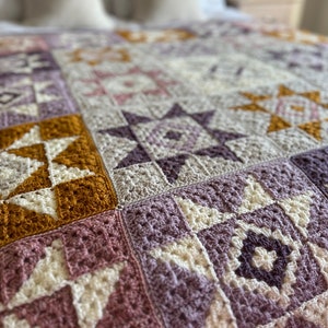Peut inclure: Une couverture en crochet avec un motif patchwork compos&eacute; de carr&eacute;s dans des tons de violet, rose, blanc et jaune moutarde.