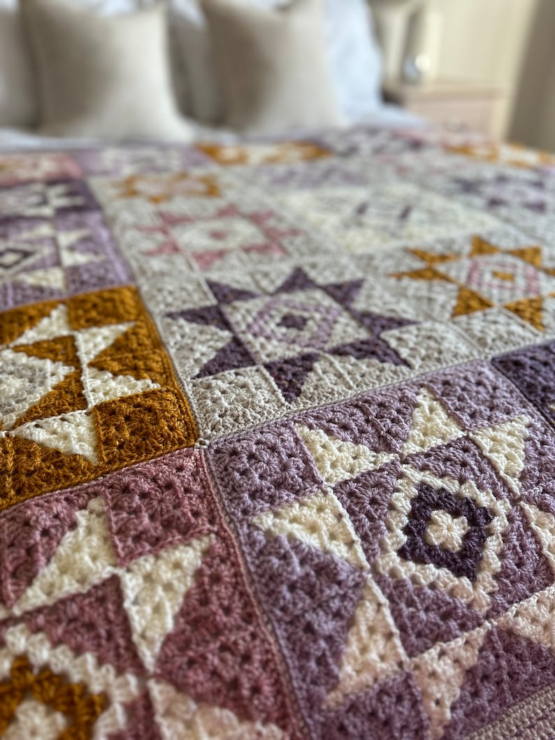 Peut inclure: Une couverture en crochet avec un motif patchwork compos&eacute; de carr&eacute;s dans des tons de violet, rose, blanc et jaune moutarde.