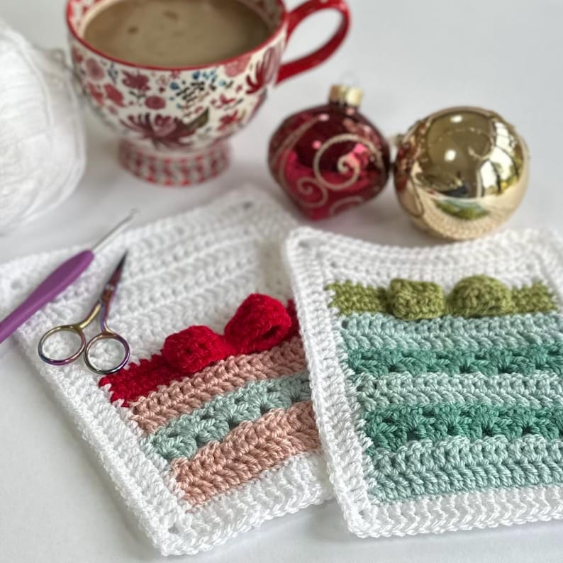 May include: Two crocheted squares, one with red, pink, and green stripes and a red bow, the other with green and white stripes. The squares are on a white surface with a cup of coffee, a red ornament, and a gold ornament.