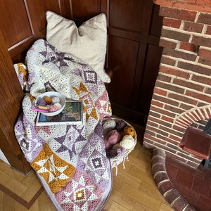 Peut inclure: Une couverture au crochet avec un motif patchwork dans des tons de violet, rose, blanc et jaune. La couverture est drap&eacute; sur une chaise avec un coussin blanc et un livre intitul&eacute; "The Lost Flock" sur le dessus. Il y a un panier de laine &agrave; c&ocirc;t&eacute; de la chaise.