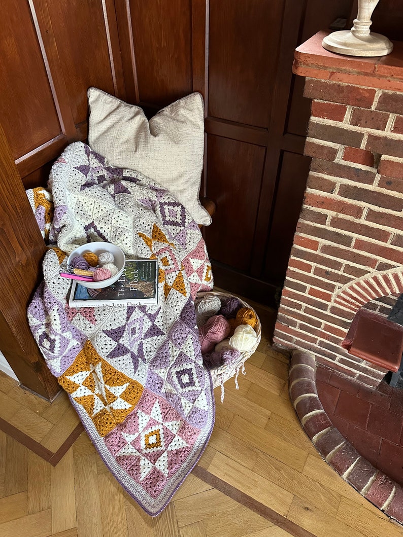 Peut inclure: Une couverture au crochet avec un motif patchwork dans des tons de violet, rose, blanc et jaune. La couverture est drap&eacute; sur une chaise avec un coussin blanc et un livre intitul&eacute; "The Lost Flock" sur le dessus. Il y a un panier de laine &agrave; c&ocirc;t&eacute; de la chaise.