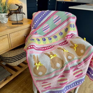 May include: A colourful, handmade crochet blanket with an Easter theme. The blanket features pastel shades of pink, purple, green, and yellow. It has patterns of Easter eggs, bunnies with yellow bows, and floral accents. The blanket is draped over a white chair.