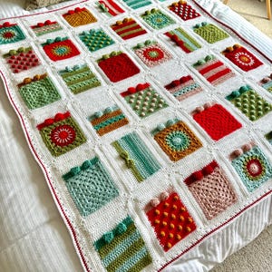 May include: A white crocheted blanket with a red border. The blanket is made up of squares with different patterns and colors, including red, green, yellow, and brown. The squares are arranged in a grid pattern.