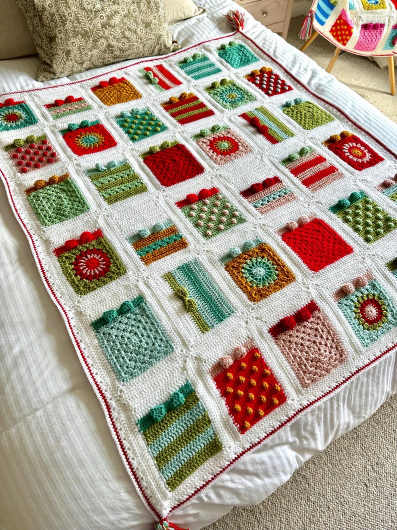 May include: A white crocheted blanket with a red border. The blanket is made up of squares with different patterns and colors, including red, green, yellow, and brown. The squares are arranged in a grid pattern.