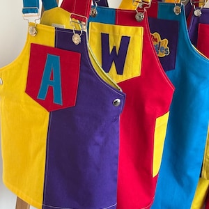 May include: A set of colourful dungarees featuring adjustable metal buckles and straps. The front displays a pocket with a large letter A or W, while another garment shows a yellow embroidered patch with the text The Wiggles. The fabric is colour-blocked in bright yellow, red, blue, and purple.