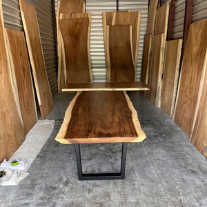 May include: A large wooden table with a live edge top and black metal legs. The table is surrounded by stacks of wood slabs in a warehouse setting.