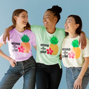 May include: Three women wearing t-shirts with a pineapple graphic and the text "cling to what is good". The shirts are in pink, light green, and cream.