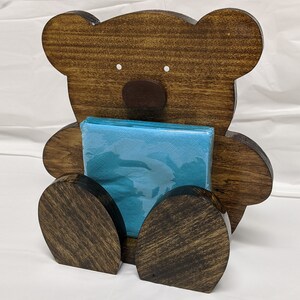 May include: A wooden bear-shaped napkin holder, stained dark brown. The bear features a brown nose and white eyes, and holds a stack of blue napkins. The holder is designed to sit upright on a table or counter.