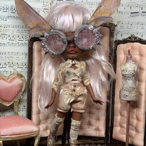 May include: A custom doll with a steampunk aesthetic, wearing goggles, fairy wings, and a vintage-inspired outfit. The doll has long, light pink hair and is posed in front of a pink backdrop. The doll's boots have skull details. The text "BLACK ORCHID DOLLS" is visible.