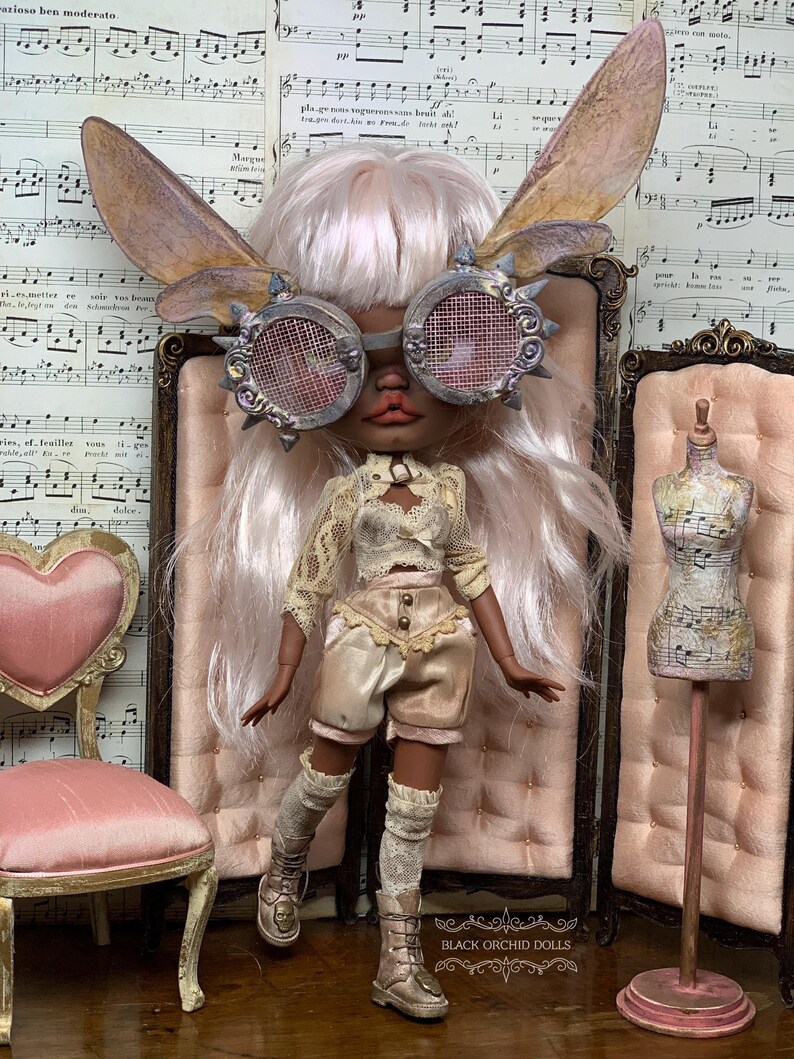 May include: A custom doll with a steampunk aesthetic, wearing goggles, fairy wings, and a vintage-inspired outfit. The doll has long, light pink hair and is posed in front of a pink backdrop. The doll's boots have skull details. The text "BLACK ORCHID DOLLS" is visible.